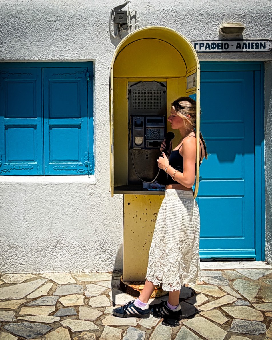 A vintage yellow phone booth against a whitewashed Greek wall with bright blue shutters